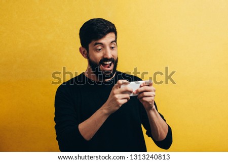 Portrait of a cheerful young man playing games on mobile phone isolated over yellow background