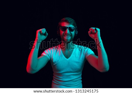 Enjoying his favorite music. Happy young stylish man in sunglasses with headphones listening sound and smiling while standing against blue neon background