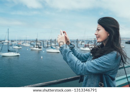 side view of young asian woman tourist taking photo of yacht with smart phone in front seat in Old Fisherman's Wharf monterey. smiling lady traveler holding cellphone standing on beach bay near boats
