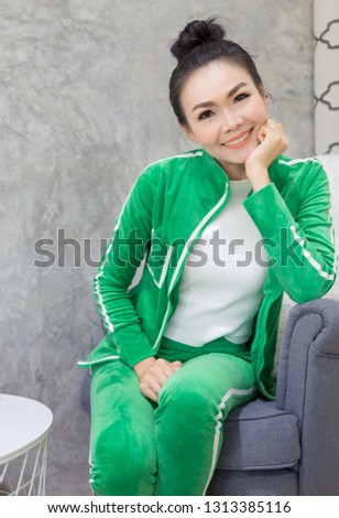 smart asian femalerelax on gray sofa, she sitting and smile, she rest chin on her hand, she wear green costume and feeling confident