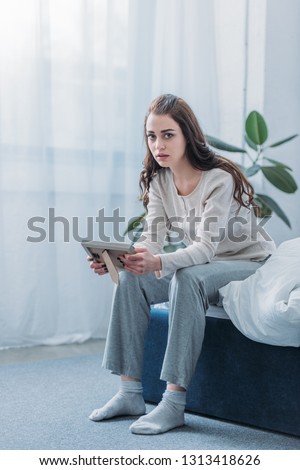 grieving woman sitting on bed, looking at camera and holding picture frame at home