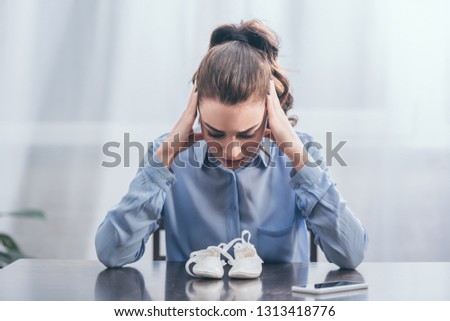 sad woman in blue blouse sitting at wooden table with smartphone and looking at white baby shoes at home, grieving disorder concept
