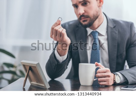 selective focus of sad man in gray suit sitting at wooden table and looking at ring in room, grieving disorder concept