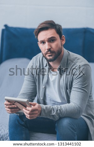 sad man sitting on bed, looking at camera and holding photo frame at home