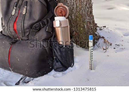 Open golden metal thermos with drink in photo camera backpack pocket on halt in winter hike under the tree trunk and Mercury thermometer in the snow showing negative temperature Celsius. Close up