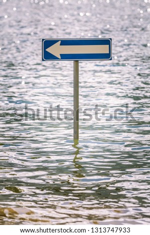 Billboard road arrow to the left partially submerged by flood water. A road sign lies in the middle of a lake of water.
