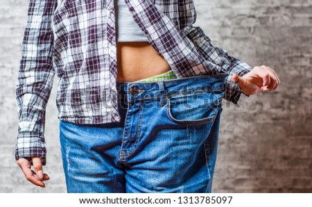 Diet concept and weight loss. Woman shows her weight loss by wearing an old jeans on gray wall background