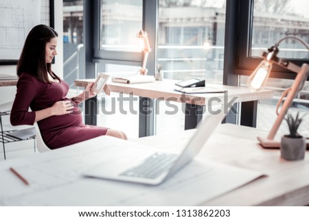 Baby bump. Appealing young dark-haired woman feeling sentimental while holding her baby bump in spacious living room