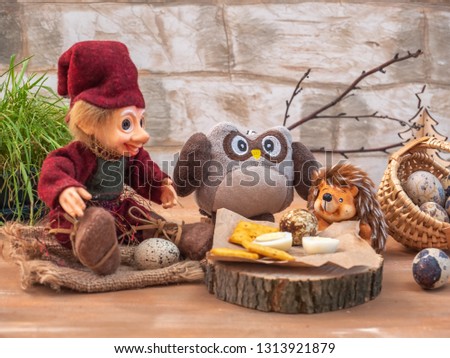 Dreamlike card with toy characters. Three friends Woodman, an owl and a hedgehog dine, they eat eggs and cracker cookies on the forest hemp.
