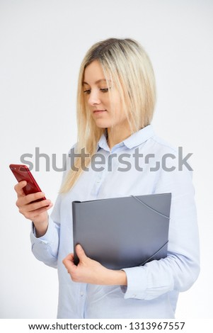 Young cute blonde caucasian woman stands sideways and looks at her red cell phone holding document folder in the office with a white background.