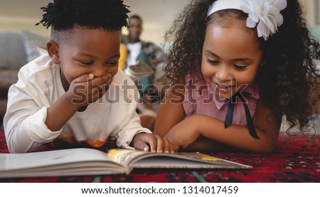 Front view of cute African American sibling lying on floor and reading a storybook in a comfortable home. Social distancing and self isolation in quarantine lockdown for Coronavirus Covid19 Royalty-Free Stock Photo #1314017459