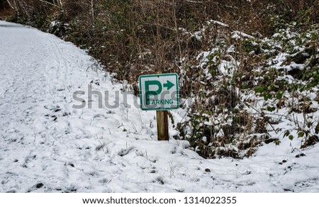 A green and white sign in the snow that says "Parking" with a large P and an arrow pointing to the right