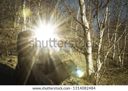 A hooded figure holding a mirror reflecting the sun, with sun flare into the camera. On a sunny winters day in a woodland.