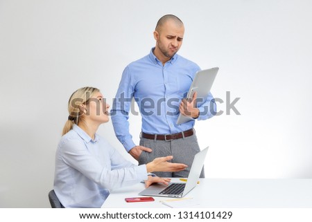 Blonde caucasian woman points her work on the screen of an aluminum laptop to her male colleague with aluminum notebook computer and cell phone. Man doesn't like it