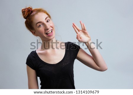 Geek saluting concept. Young red headed girl in casual black shirt welcomes fans of fiction films Vulcan greeting on grey background copy space