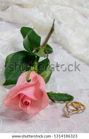 narrow wedding band and diamond engagement ring with pink rose on a white lace background vertical