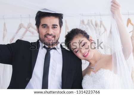 Happiness couple newlywed beautiful Asian bride and mix race or foreigner groom hold hand lie her head on groom shoulder in the wedding dress fitting room background, marriage with foreigner concept