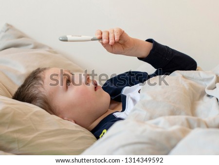 Pre-school sick boy in pyjama lying on pillow in bed and looking at digital thermometer. Ill boy is measuring body temperature and doesn’t feel well. - Image