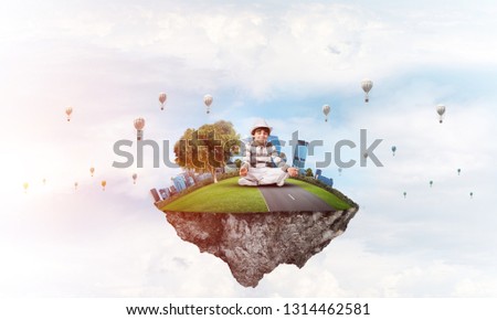 Young little boy keeping eyes closed and looking concentrated while meditating on flying island in the air with cloudy skyscape and flying aerostats on background. 3D rendering.