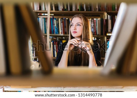 Beautiful young woman wearing glasses in the library among the bookshelves holding glasses and looking 