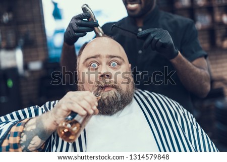 Portrait of silly and funny man sitting in armchair indoor barbershop with beverage in hands. Cropped barber combing and cutting hair of customer