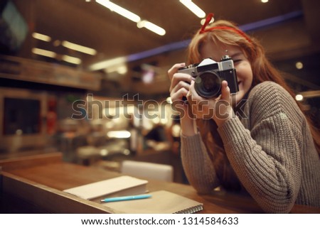 Charming girl in headband photographing while sitting at the table with book, spiral notebook and pen in cafe. Copy space in left side