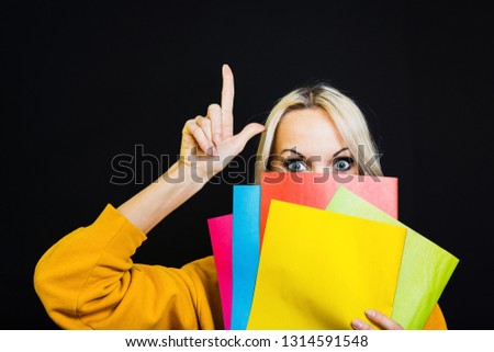 The image of a student girl, hiding her face behind books, raises her index finger up with surprised eyes, stands on a black background.  The concept of school, University.