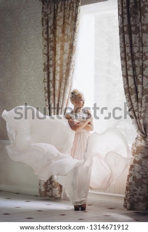 Cute blonde girl in the image of the bride stands near the window in a flying bright dress. wedding photo. gentle calm image. Small Noise