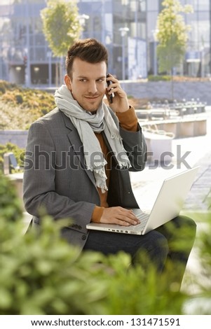 Young businessman sitting in citypark, talking on mobilephone, using laptop computer.