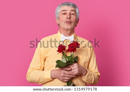 Isolated shot of attractive wrinkled grey haired man holds red roses, dressed in yellow elegant shirt and bowtie, keeps lips folded, going to kiss someone. Grandfather recieves flowers on birthday