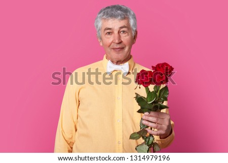 Elegant romantic mature gentleman in formal shirt with bowtie holds red roses, ready for date or birthday, looks directly at camears with satisfied expression, isolated over pink background.