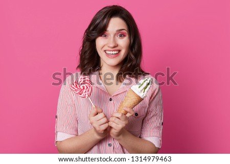 Photo of attractive lovely young woman with brown hair, holds tasty candy and ice cream, wears striped shirt, looks at camera with satisfaction, being sweet tooth, isolated over pink background