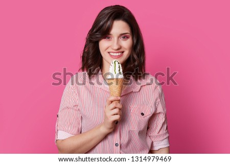 Picture of smiling young woman with pleased expression, toothy smile, holds tasty ice cream, enjoys leisure, dressed in fashionable shirt, models over rosy background, appetizes favorite dessert