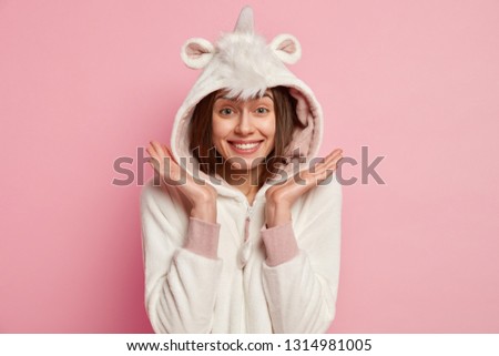 Headshot of good looking woman with pleased facial expression, spreads palms near face, has toothy smile, wears white kigurumi costume with hoody, isolated over pink background. Sleeping concept
