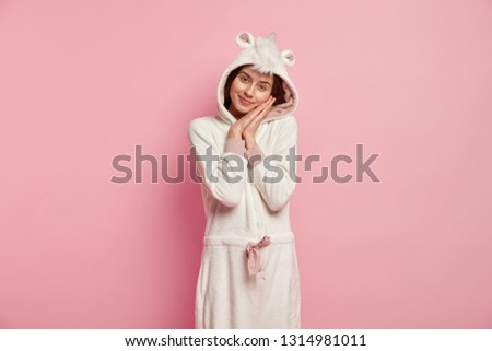 Restful girl leans at hands, enjoys good sleep, wears funny soft domestic outfit, looks at camera with tender look, poses over pink background, has nice rest in calm atmosphere. Bedding concept