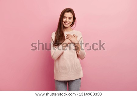 Impressed friendly pleasant looking woman keeps hands on chest, touched by compliment, smiles positively, wears jumper and jeans, models over pink background, looks at camera with great pleasure