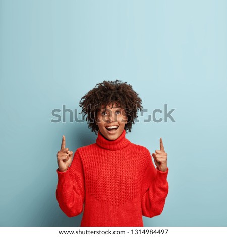 Photo of merry dark skinned woman shows direction upwards, wears red jumper, isolated over blue background with empty space for your promotional content. Upbeat pleased Afro American lady indoor