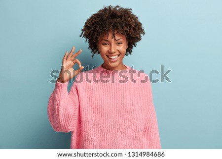 Headshot of positive dark skinned woman with crisp hair, makes okay gesture, wears loose sweater, enjoys life, isolated over blue background, says ok, confirms information. Its excellent result