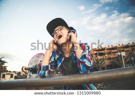 Beautiful blond child with glasses plaid shirt hat skater singing and listening to music with headphones leaning against railing with skateboard. Blue sky clouds in background. Happy young boy