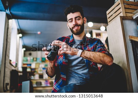 Portrait of handsome hipster photographer dressed in casual shirt smiling at camera.Cheerful young man with beard holding vintage camera and making setting on device sitting in own studio