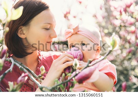 Loving mother and baby girl are smelling blooming blossom pink magnolia tree. Young beautiful mom and little daughter among garden flowers outdoors. Family on nature in Arboretum, Slovenia