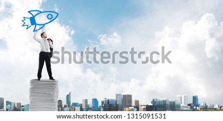 Horizontal shot of successful and young businessman in suit throwing in the air big drawn rocket while standing on paper column with modern cityscape view on background.