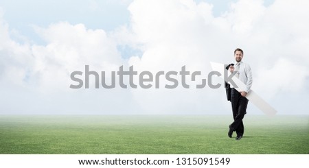 Confident and young businessman in suit holding big white arrow in hands which pointing to the side while standing on green lawn and cloudy skyscape on background.