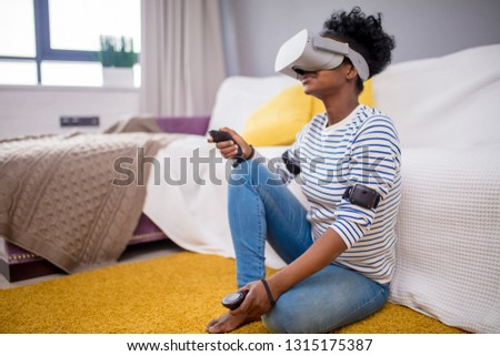Indoor shot of pretty african female gamer using augmented reality headset with modern 3d mobile app.Young female experience virtual reality with smartphone and VR glasses on her head.