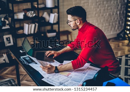 Concentrated male it developer working remotely with paper documents and modern laptop computer during time at stylish apartment, serious students in casual look checking information on website