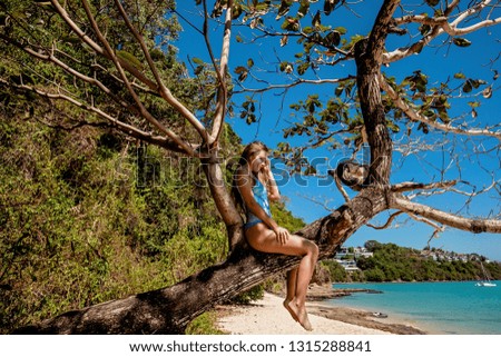 Happy young girl smiling and sitting on tropical tree at the island beach with clear water on the yachts background while the sky is blue