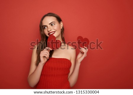Smiling beautiful woman looking aside while holding nice decorations in studio. She is keeping Valentine cards in hands and touching her face with decorations