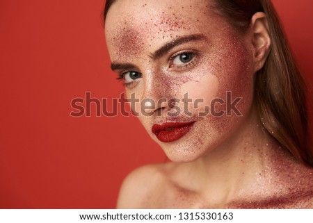 Female face covered with glossy glitter make up and empty heart-shaped picture on face. She is looking at camera while posing in studio indoors. Close up