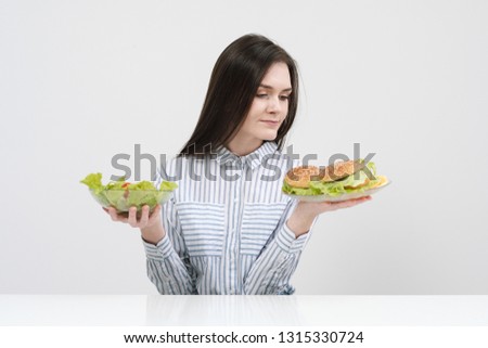 Slender brunette girl on a white background chooses between a plate of fast food and hamburgers and healthy food salad. Dieting concept.