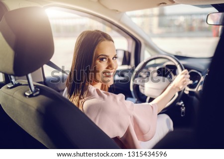 Pretty Caucasian brunette driving car and looking at camera. Picture taken from back seat.